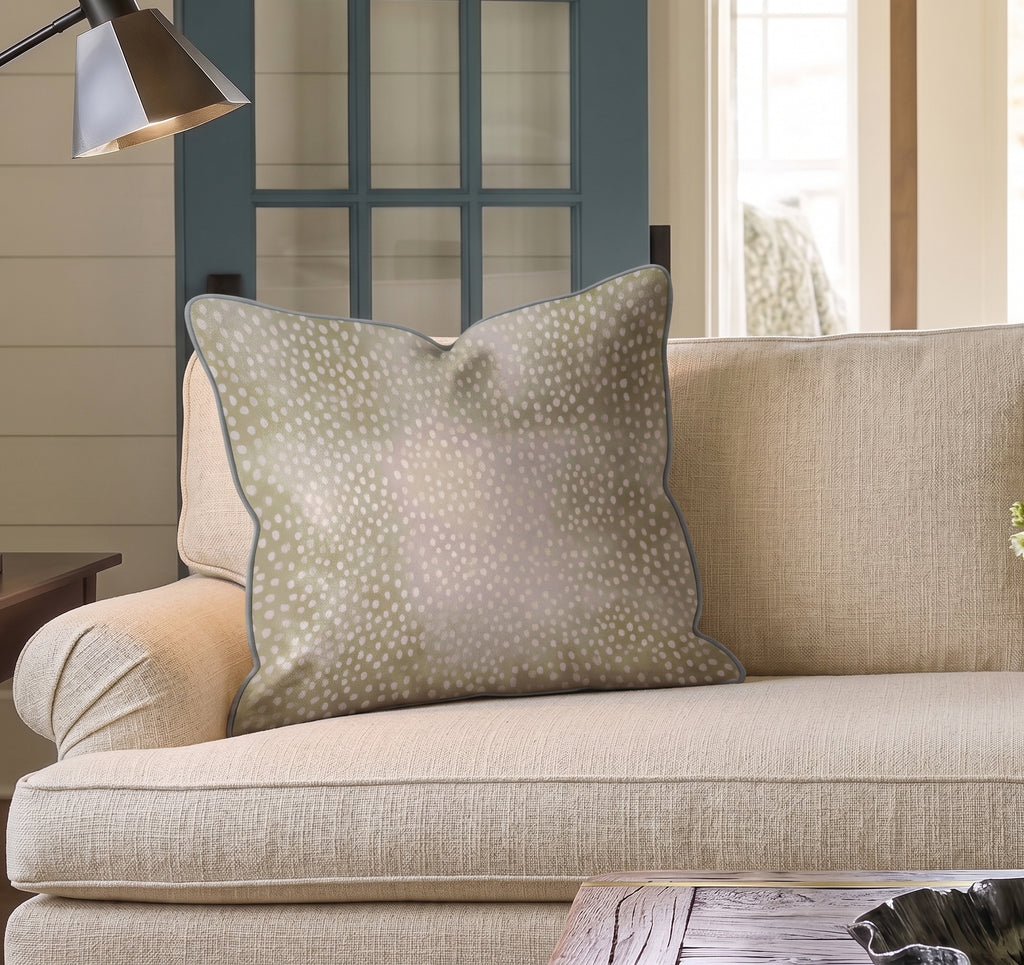 A beige couch with a large, square Hillcroft Home Dotted Powder Fawn Throw Pillow, crafted from a linen blend and made in the USA, sits in a cozy living room with a blue door and a lamp nearby.
