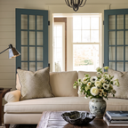 A beige sofa with patterned pillows—including the Hillcroft Home Dotted Brown Fawn Throw Pillow—sits in front of blue French doors. A vase of white flowers rests on a wooden coffee table, and sunlight streams through the windows in the background.