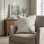 A beige linen blend armchair topped with the Hillcroft Home Hillcroft x Lemon Park - Tangled Throw Pillow sits beside a wooden side table arranged with books, a framed photo, and a vase of dried branches in a softly lit, elegant room.